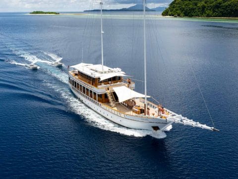 Fenides luxury motor yacht underway from above on deep blue water with tender following and lush green tropical Indonesian islands behind