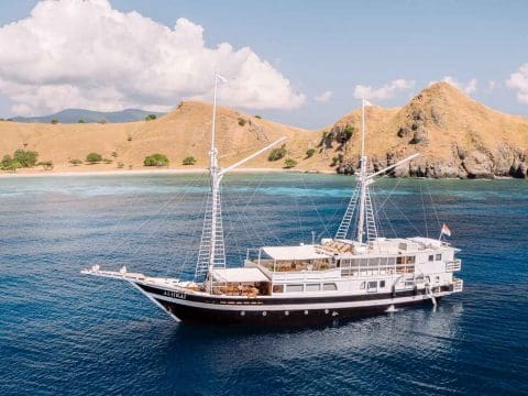 Alii Kai luxury phinisi liveaboard with dark hull anchored in blue Komodo waters with iconic dry volcanic golden hills and turquoise bay behind