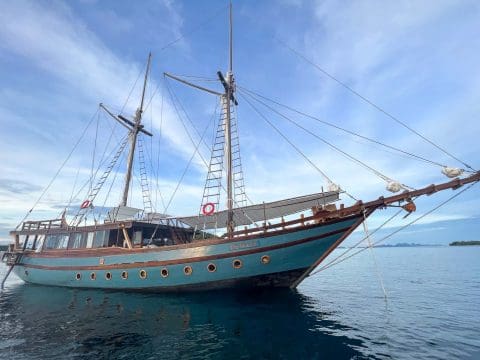 Dewata traditional Indonesian phinisi yacht with distinctive teal hull and twin masts anchored in calm water with tropical islands behind