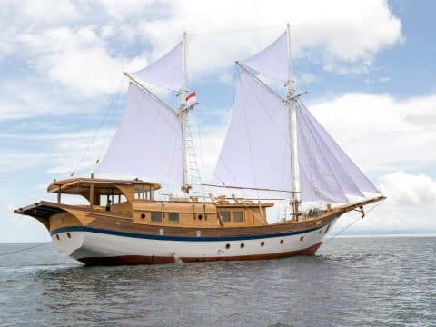 Leyla luxury Indonesian phinisi charter yacht under white sails with warm wood superstructure, white hull and Indonesian flag on calm open sea