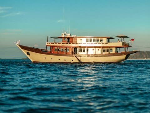 Mischief classic luxury motor yacht with cream hull and dark wood cabin anchored in calm blue Indonesian waters with green hills behind
