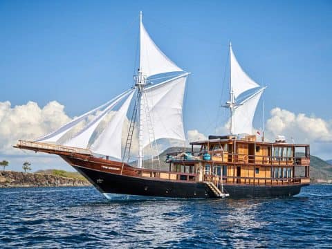 The Oracle luxury phinisi liveaboard under full white sails with dark hull and wood cabin cruising Komodo National Park waters with rocky islands behind