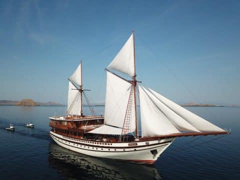 Prana luxury Indonesian phinisi liveaboard with full white sails deployed anchored in Komodo National Park with dry volcanic hills behind