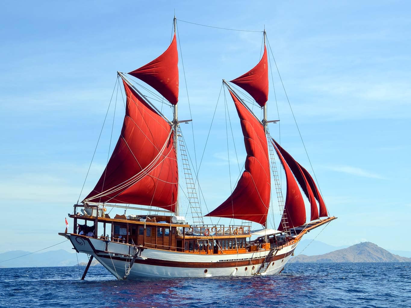 Tiare luxury Indonesian phinisi sailing yacht under full deep red crimson sails with wooden hull and volcanic island in the background
