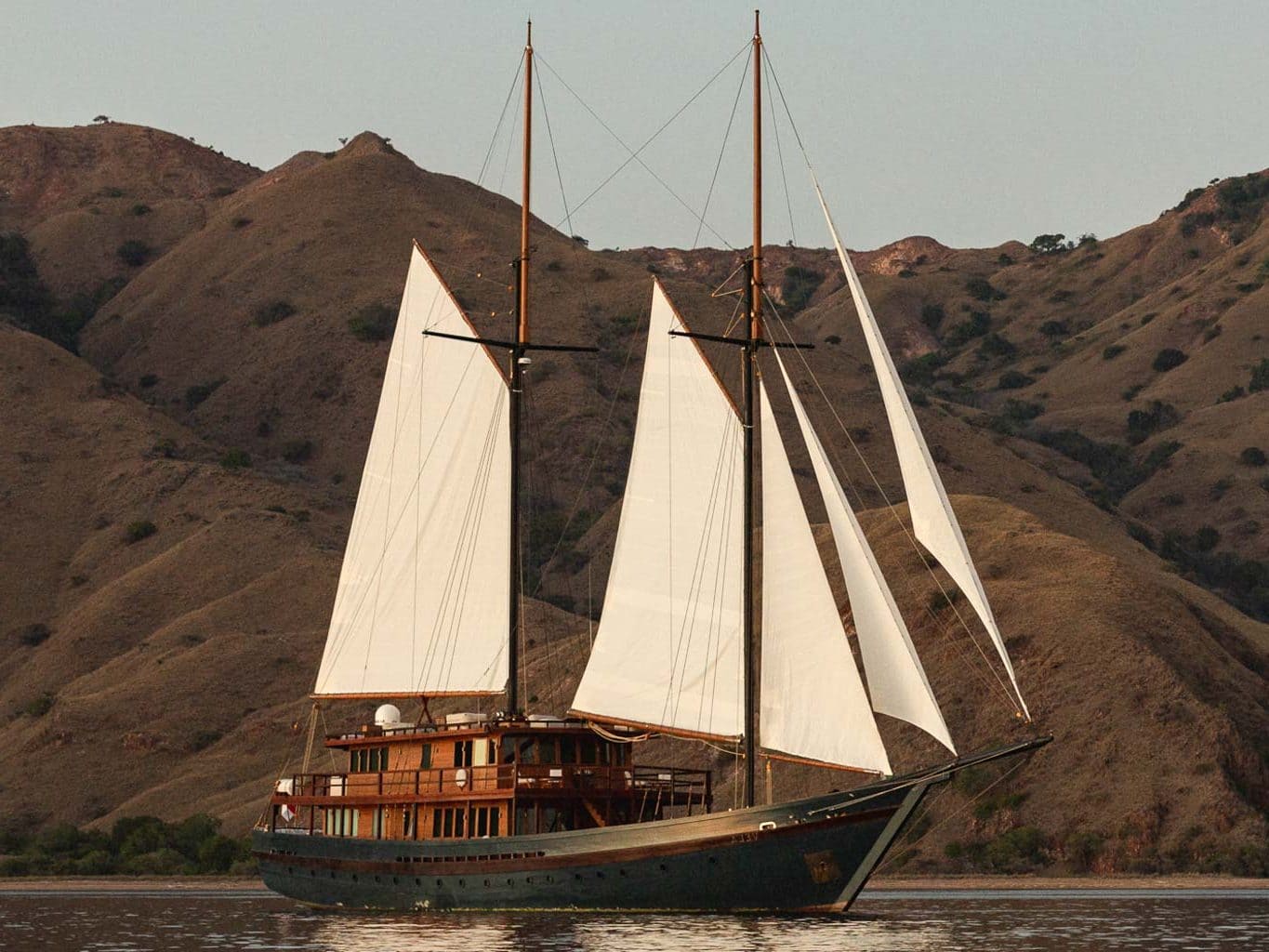 Vela liveaboard sailing under full white sails with three masts deployed against dramatic rocky Komodo National Park hillsides
