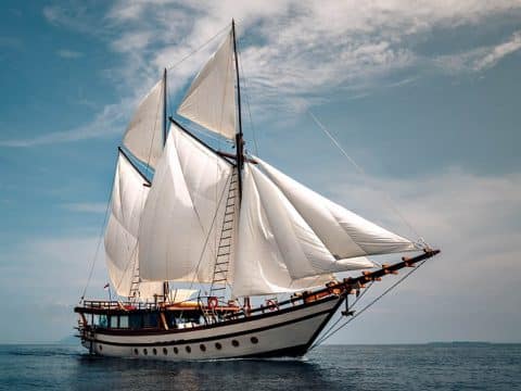 Upward view of Senja phinisi liveaboard white sails fully billowing against blue sky and clouds