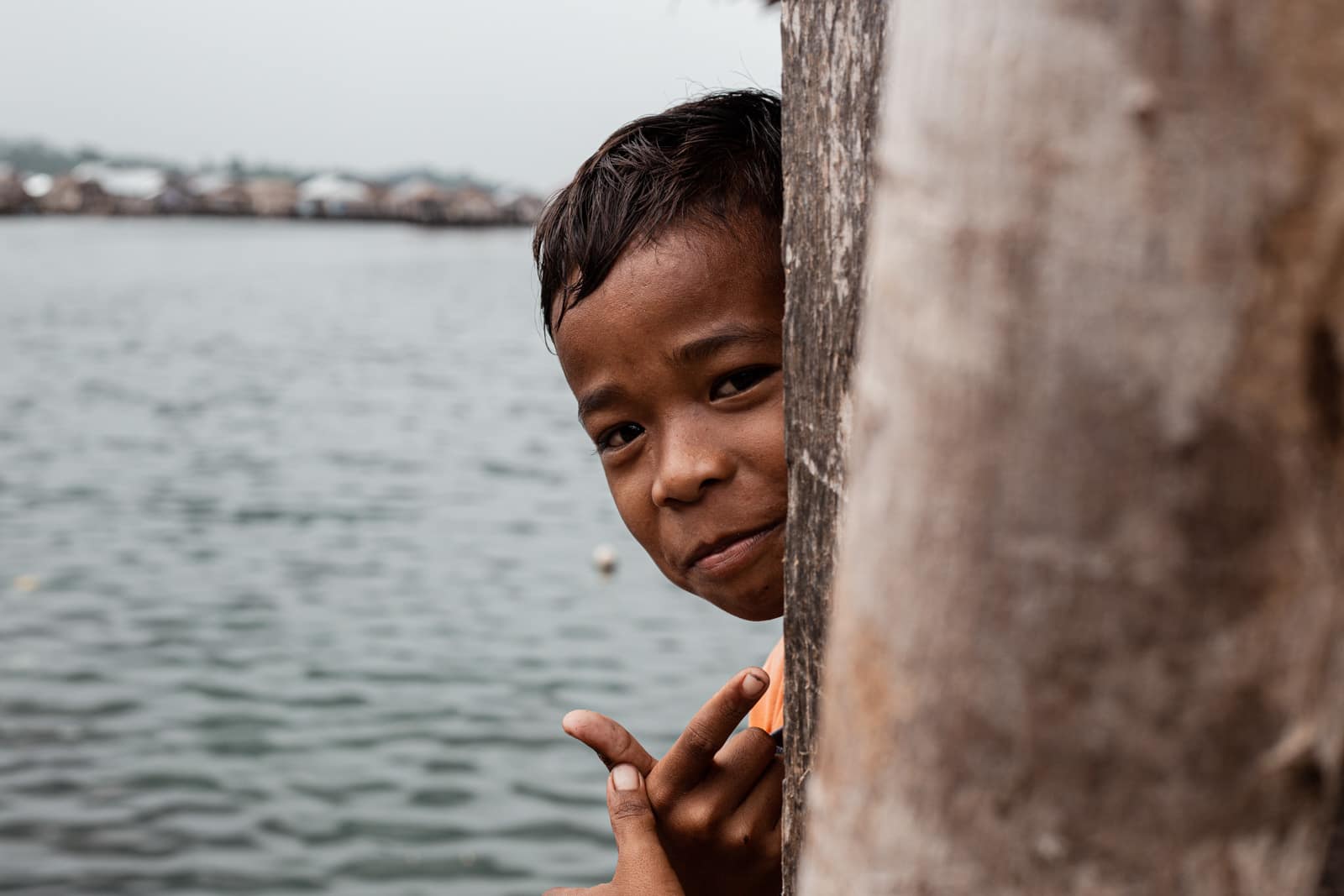 Young Bajau child peeking around a wooden post and making a peace sign, Bajau village in the background