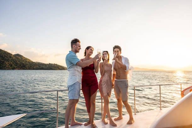Group of four friends toasting champagne glasses at golden hour sunset on a yacht with a tropical island in the background